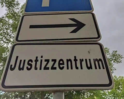 Straßenschild mit Pfeil und der Aufschrift „Justizzentrum“ vor Bäumen und Himmel.