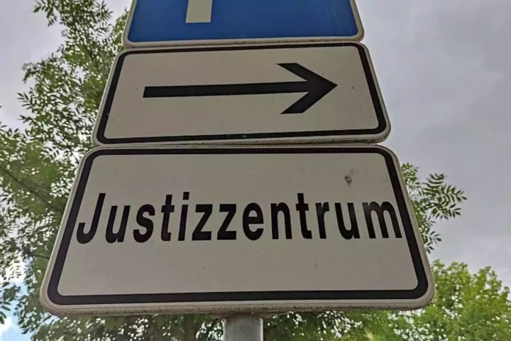 Straßenschild mit Pfeil und der Aufschrift „Justizzentrum“ vor Bäumen und Himmel.