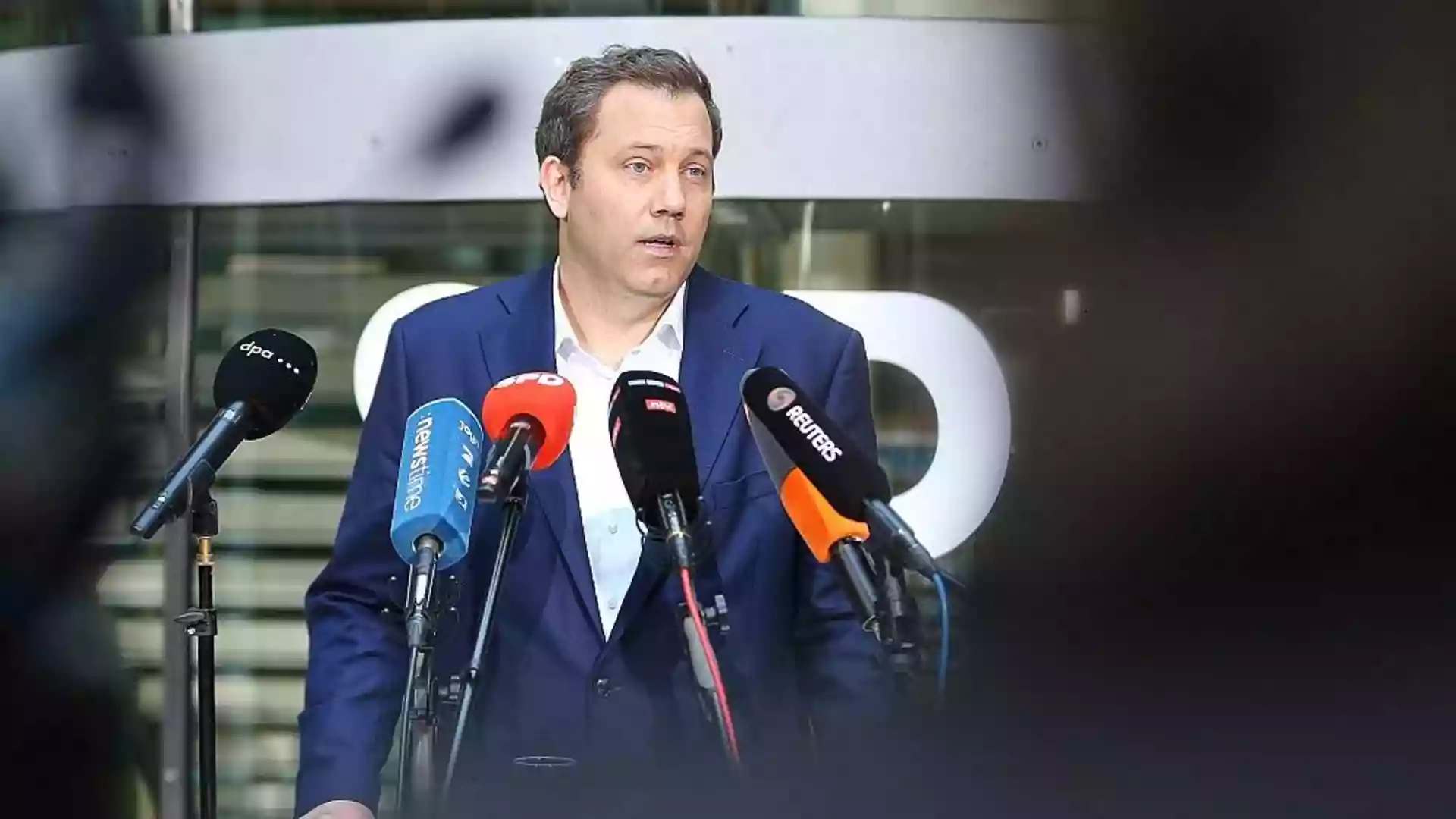 Mann im blauen Anzug spricht vor mehreren Mikrofonen bei einer Pressekonferenz.
