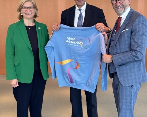 Friedrich Merz steht mit zwei weiteren Personen und hält ein hellblaues Team-Saarland-Shirt.