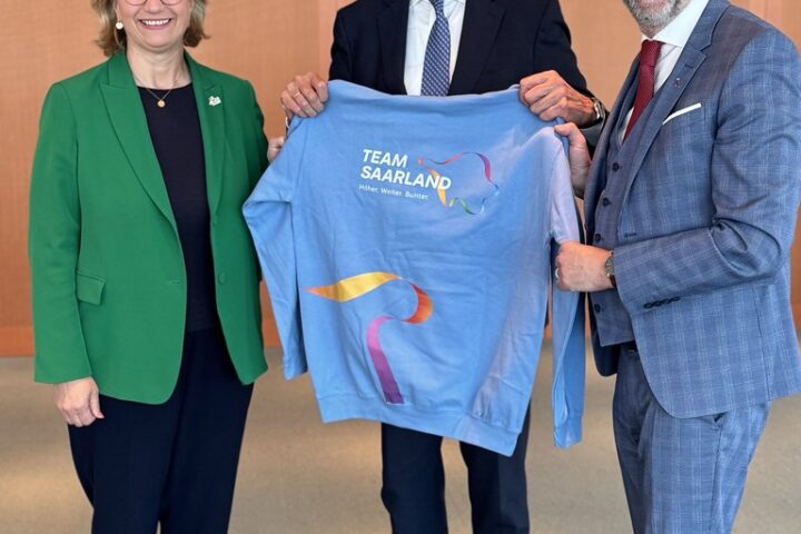 Friedrich Merz steht mit zwei weiteren Personen und hält ein hellblaues Team-Saarland-Shirt.