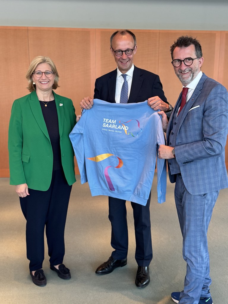 Friedrich Merz steht mit zwei weiteren Personen und hält ein hellblaues Team-Saarland-Shirt.