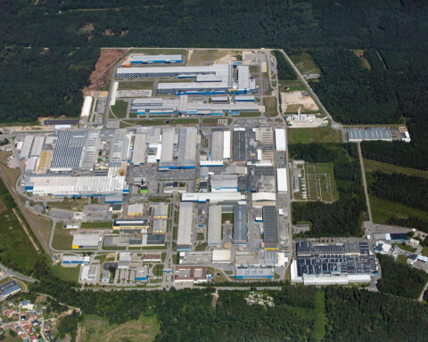 Luftaufnahme eines großen Industrie- und Werkgeländes mit mehreren Produktionshallen und Straßen inmitten von Waldgebiet.