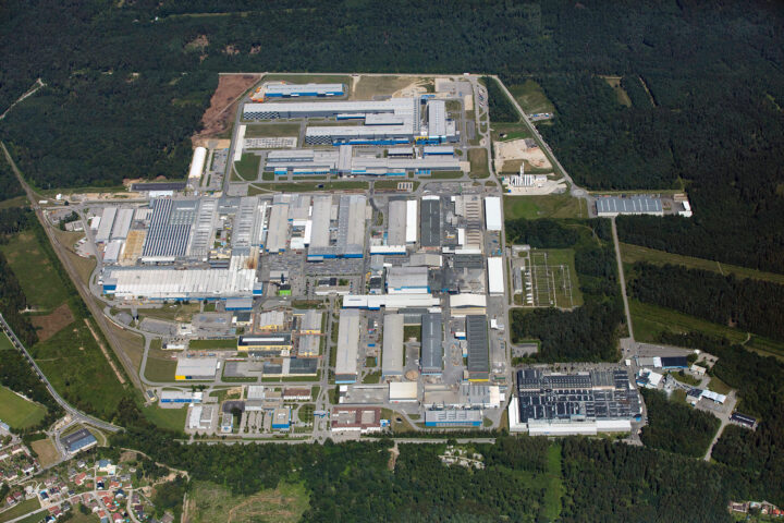 Luftaufnahme eines großen Industrie- und Werkgeländes mit mehreren Produktionshallen und Straßen inmitten von Waldgebiet.