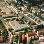 Luftaufnahme eines großen Industrie- und Werksgeländes mit mehreren Hallen und Gebäuden in einer Wohnumgebung.