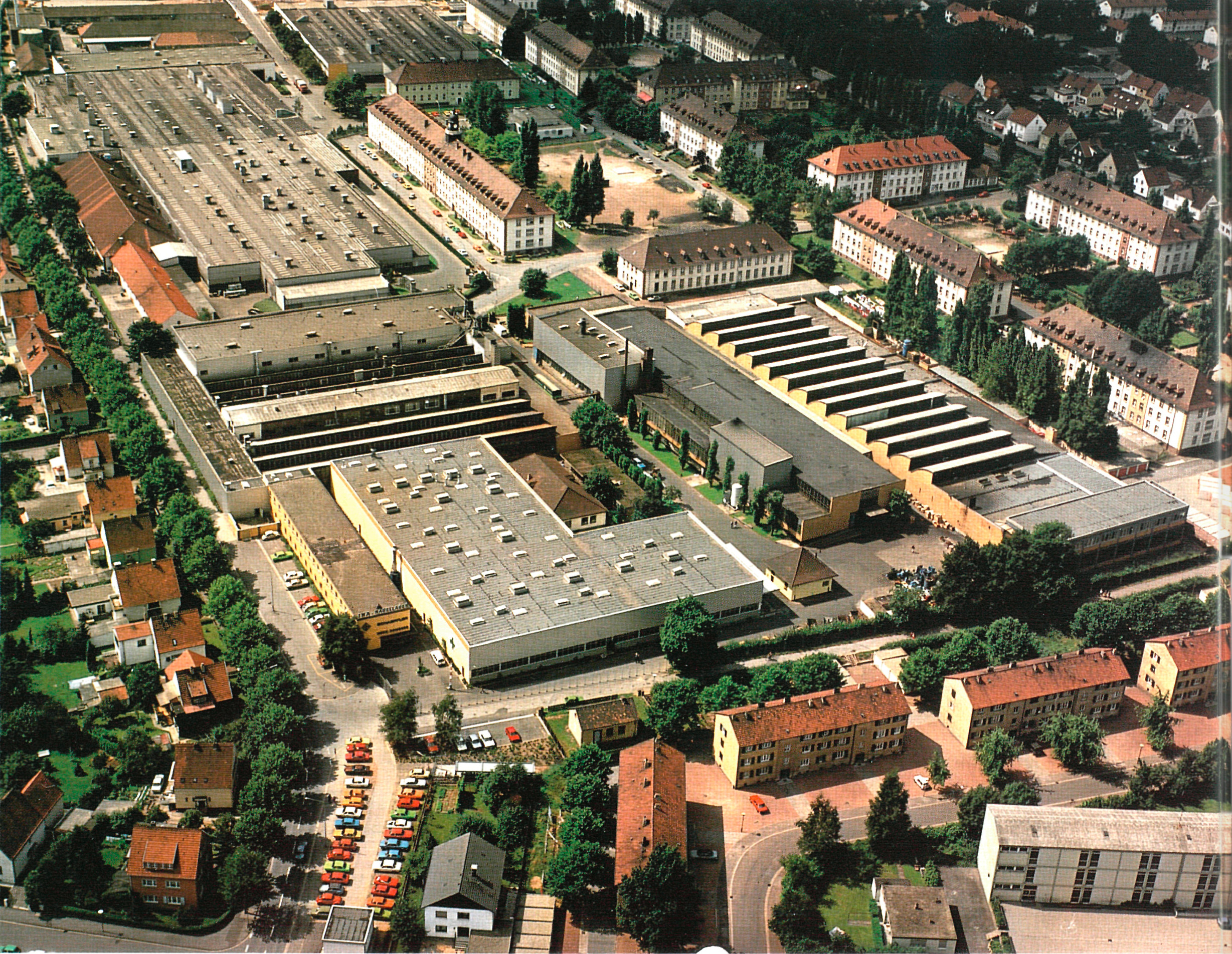 Luftaufnahme eines großen Industrie- und Werksgeländes mit mehreren Hallen und Gebäuden in einer Wohnumgebung.