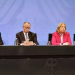 Vier Politiker sitzen an einem Konferenztisch vor blauem Hintergrund mit Bundesadler.
