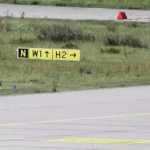 Rollbahnmarkierung auf einer Flugplatzfläche mit gelbem Schild und Grasstreifen.