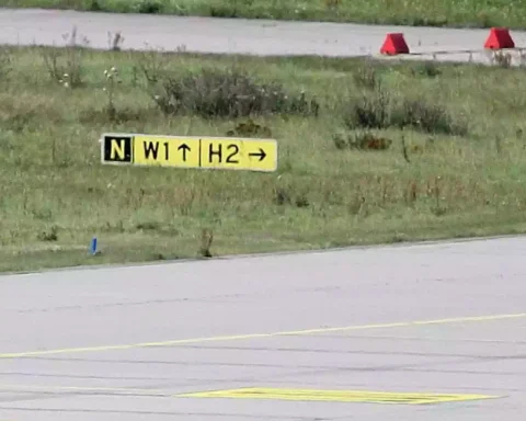 Rollbahnmarkierung auf einer Flugplatzfläche mit gelbem Schild und Grasstreifen.