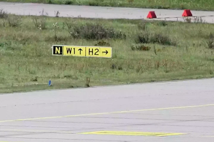 Rollbahnmarkierung auf einer Flugplatzfläche mit gelbem Schild und Grasstreifen.