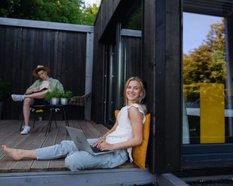 Zwei Personen sitzen auf der Holzterrasse eines modernen Tiny Houses, eine Frau mit Laptop im Vordergrund.