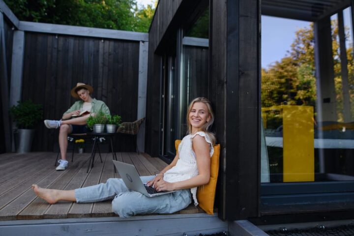 Zwei Personen sitzen auf der Holzterrasse eines modernen Tiny Houses, eine Frau mit Laptop im Vordergrund.