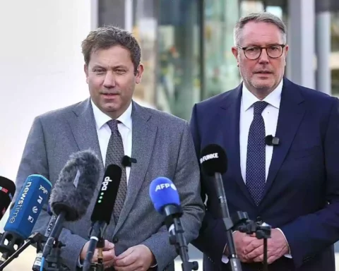 Alexander Schweitzer steht bei einer Pressekonferenz vor mehreren Mikrofonen neben einem weiteren Mann im Anzug.