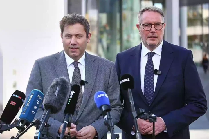 Alexander Schweitzer steht bei einer Pressekonferenz vor mehreren Mikrofonen neben einem weiteren Mann im Anzug.