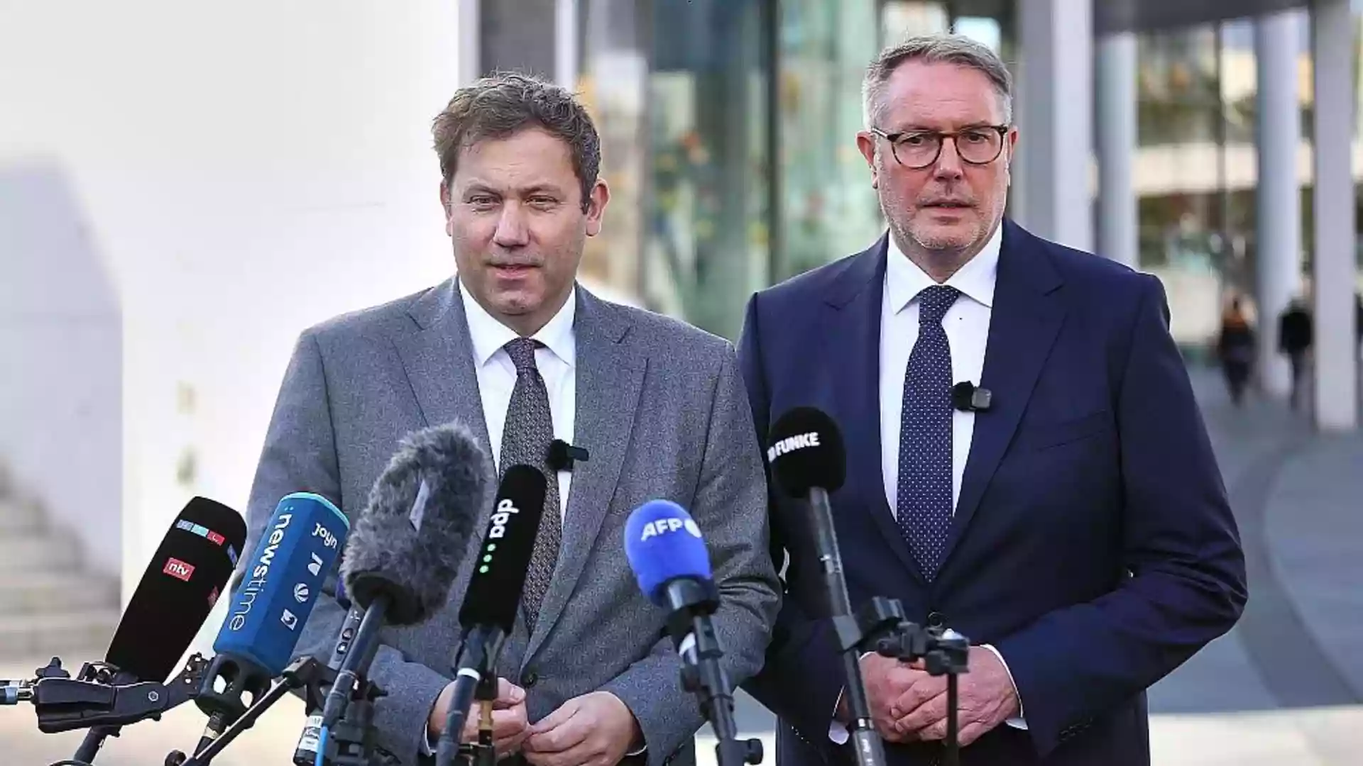 Alexander Schweitzer steht bei einer Pressekonferenz vor mehreren Mikrofonen neben einem weiteren Mann im Anzug.