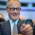Alexander Schweitzer lächelt bei einer Veranstaltung im Innenraum, umgeben von weiteren Personen.