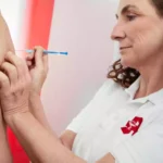 Eine Frau in weißem Poloshirt setzt einer Person eine Injektion in den Oberarm.