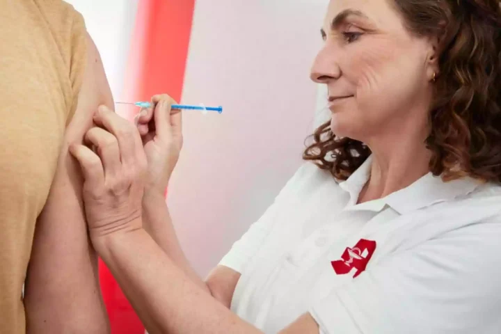 Eine Frau in weißem Poloshirt setzt einer Person eine Injektion in den Oberarm.