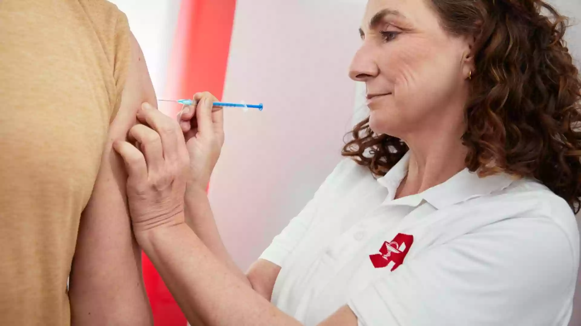 Eine Frau in weißem Poloshirt setzt einer Person eine Injektion in den Oberarm.