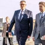 Alexander Schweitzer geht mit weiteren Personen im Freien entlang einer Uferpromenade.