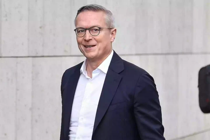 Karsten Wildberger steht vor einer hellen Steinwand und lächelt in die Kamera.