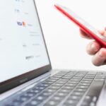 Hand hält ein Smartphone vor einem Laptop mit Anzeige von Online-Zahlungsoptionen.