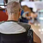 Ein Mann sitzt im Rollstuhl in einem Pflegeheim oder Aufenthaltsraum, im Hintergrund sind unscharf weitere Personen und Tische zu sehen.