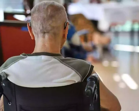 Ein Mann sitzt im Rollstuhl in einem Pflegeheim oder Aufenthaltsraum, im Hintergrund sind unscharf weitere Personen und Tische zu sehen.
