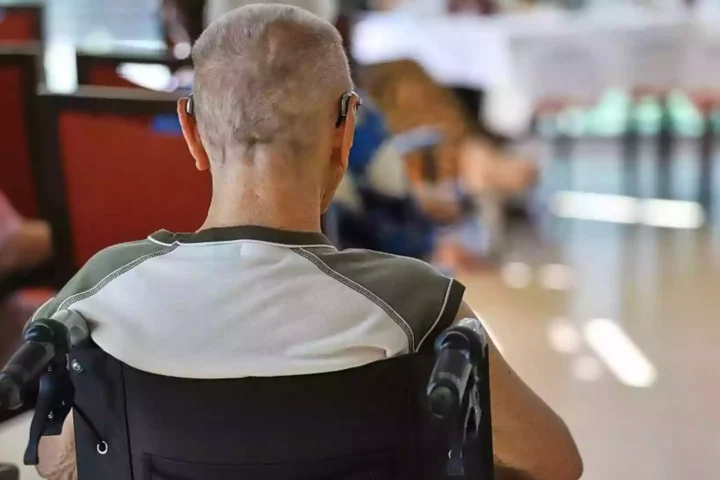 Ein Mann sitzt im Rollstuhl in einem Pflegeheim oder Aufenthaltsraum, im Hintergrund sind unscharf weitere Personen und Tische zu sehen.