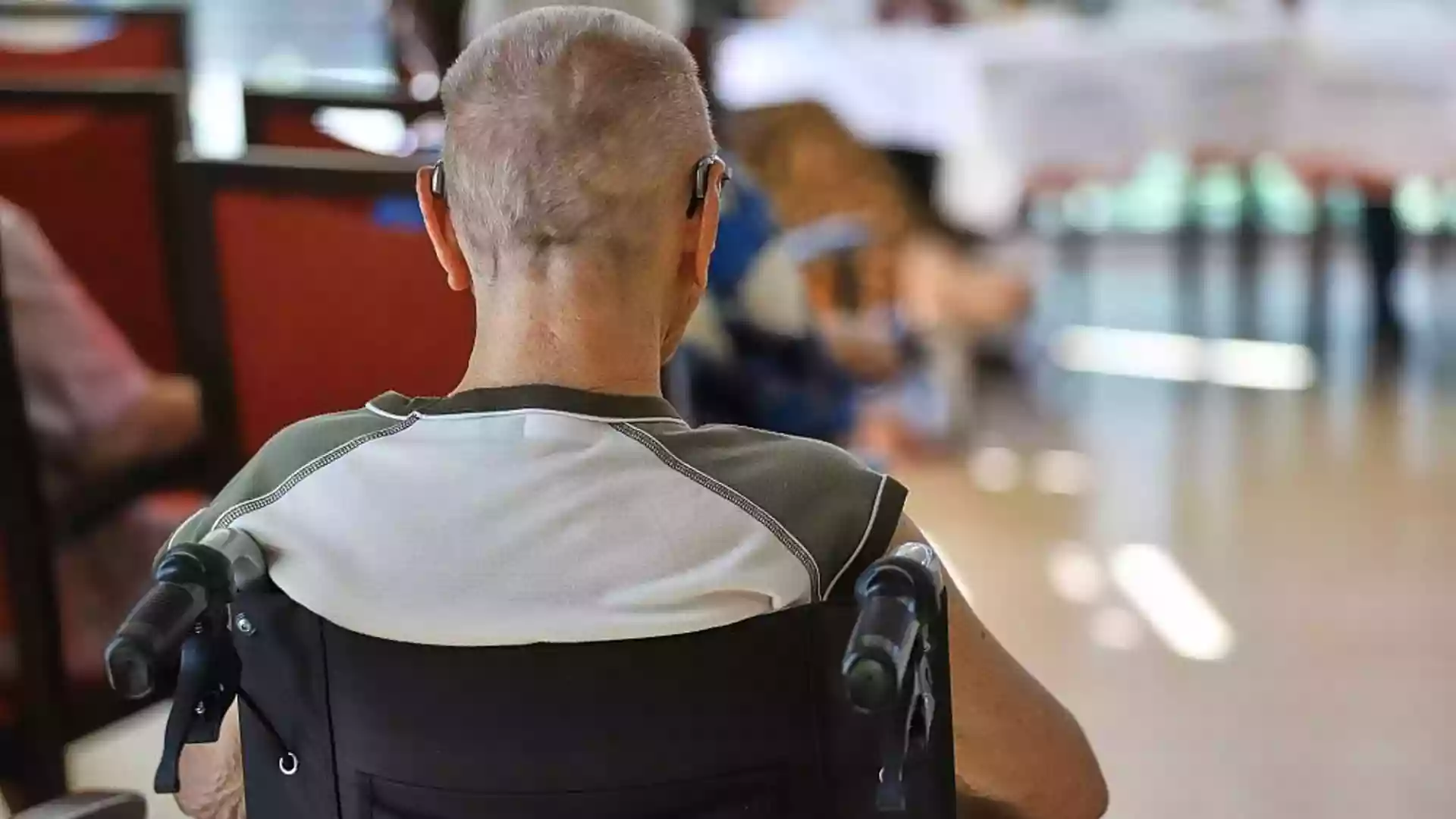 Ein Mann sitzt im Rollstuhl in einem Pflegeheim oder Aufenthaltsraum, im Hintergrund sind unscharf weitere Personen und Tische zu sehen.