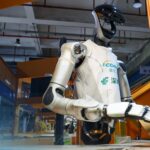 Ein humanoider Roboter greift in einer Industriehalle einen Karton.