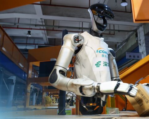 Ein humanoider Roboter greift in einer Industriehalle einen Karton.