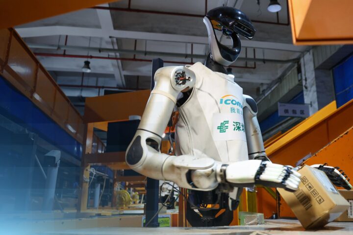 Ein humanoider Roboter greift in einer Industriehalle einen Karton.