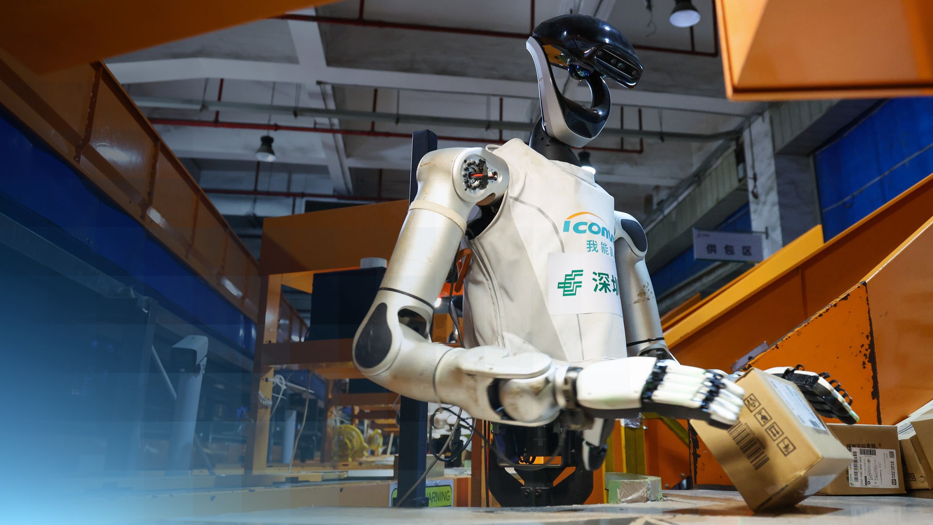 Ein humanoider Roboter greift in einer Industriehalle einen Karton.
