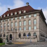 Außenansicht eines repräsentativen historischen Gebäudes mit rotem Ziegeldach unter blauem Himmel.