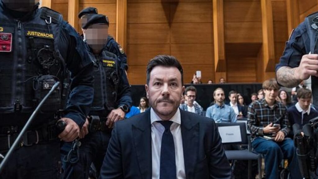 René Benko sitzt in einem Gerichtssaal, flankiert von Justizwachebeamten.