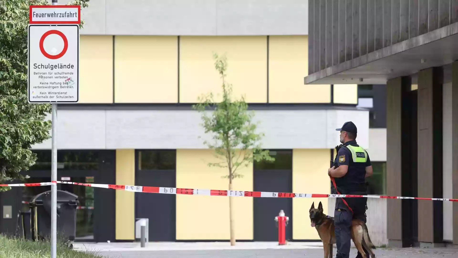 Polizist mit Diensthund steht vor abgesperrtem Schulgelände an einem Gymnasium.