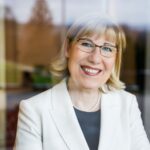 Porträt einer lächelnden Frau mit Brille und weißem Blazer vor unscharfem Hintergrund.