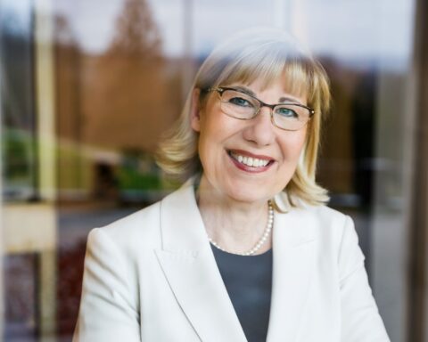 Porträt einer lächelnden Frau mit Brille und weißem Blazer vor unscharfem Hintergrund.
