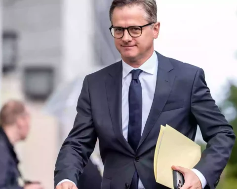 Ein Mann im dunklen Anzug mit Brille hält eine Aktenmappe in der Hand.