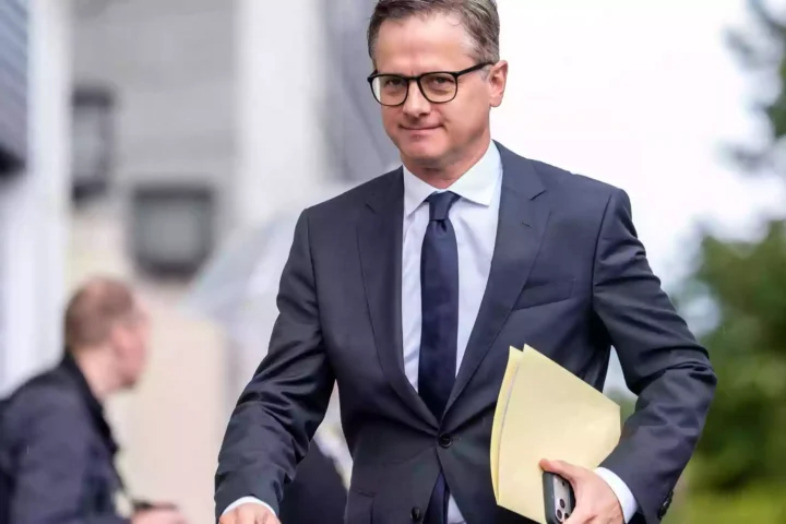 Ein Mann im dunklen Anzug mit Brille hält eine Aktenmappe in der Hand.