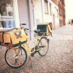 Gelbes Postfahrrad mit Transporttaschen steht auf einem Gehweg in einer Stadtstraße.