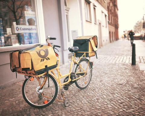 Gelbes Postfahrrad mit Transporttaschen steht auf einem Gehweg in einer Stadtstraße.