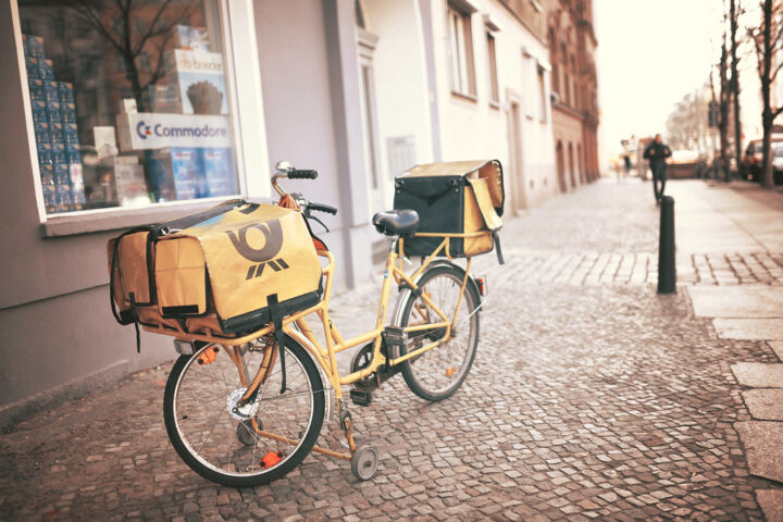 Gelbes Postfahrrad mit Transporttaschen steht auf einem Gehweg in einer Stadtstraße.