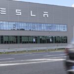 Tesla-Fabrikgebäude mit großem Tesla-Schriftzug an der Fassade, davor eine Straße und ein vorbeifahrendes Auto.