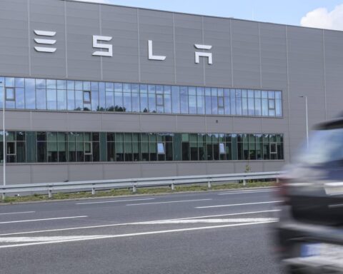 Tesla-Fabrikgebäude mit großem Tesla-Schriftzug an der Fassade, davor eine Straße und ein vorbeifahrendes Auto.
