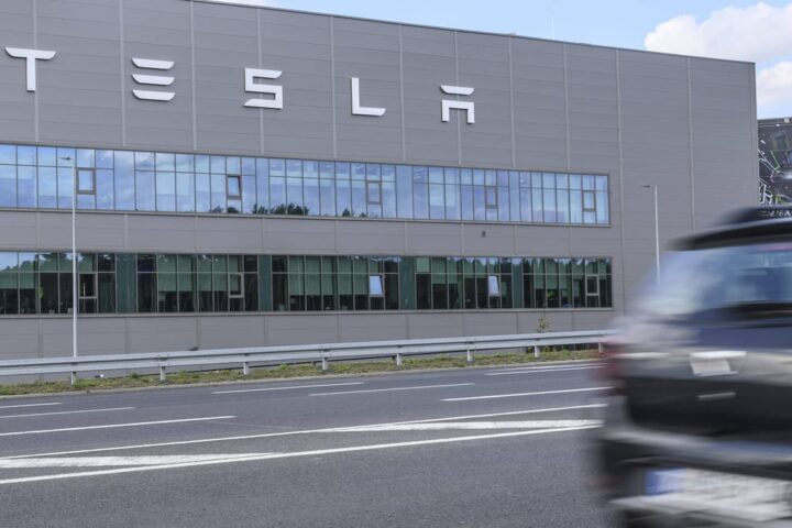 Tesla-Fabrikgebäude mit großem Tesla-Schriftzug an der Fassade, davor eine Straße und ein vorbeifahrendes Auto.