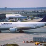 Lufthansa-Flugzeug auf dem Vorfeld am Flughafen, im Hintergrund weitere Maschinen und Abfertigungsfahrzeuge.