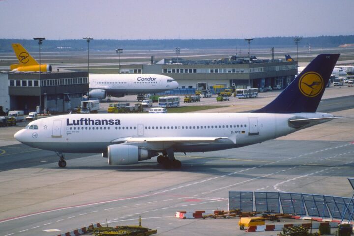 Lufthansa-Flugzeug auf dem Vorfeld am Flughafen, im Hintergrund weitere Maschinen und Abfertigungsfahrzeuge.