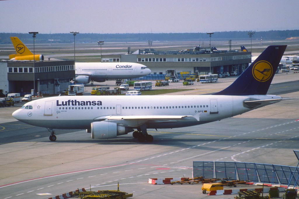 Lufthansa-Flugzeug auf dem Vorfeld am Flughafen, im Hintergrund weitere Maschinen und Abfertigungsfahrzeuge.